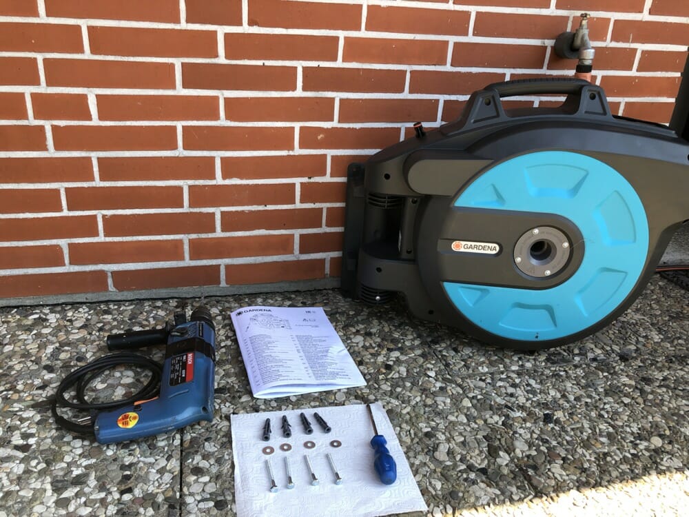 Gardena Wand-Schlauchbox vor der Montage an einer Klinkerwand, davor Bohrmaschine, Anleitung, Dübel und Schrauben